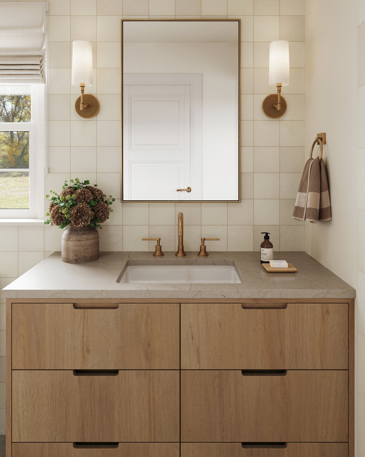 Warm neutral bathroom vanity with wood drawers, brass faucet, framed mirror, and brass wall sconces against soft eggshell tile.