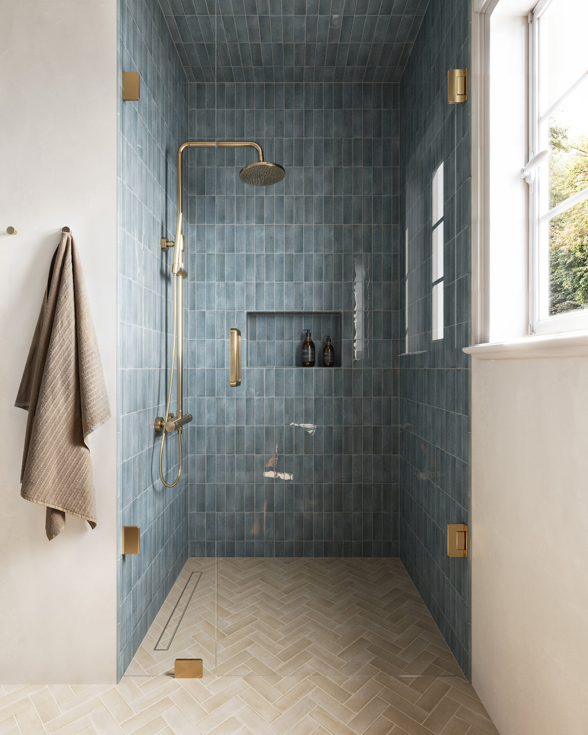 Natasha 0.5x8 glossy ceramic tile trim in Ice, accenting a serene shower clad in glossy sky-blue tiles with brass fixtures, warm beige herringbone floor tiles, and natural light for a sophisticated, spa-like ambiance.