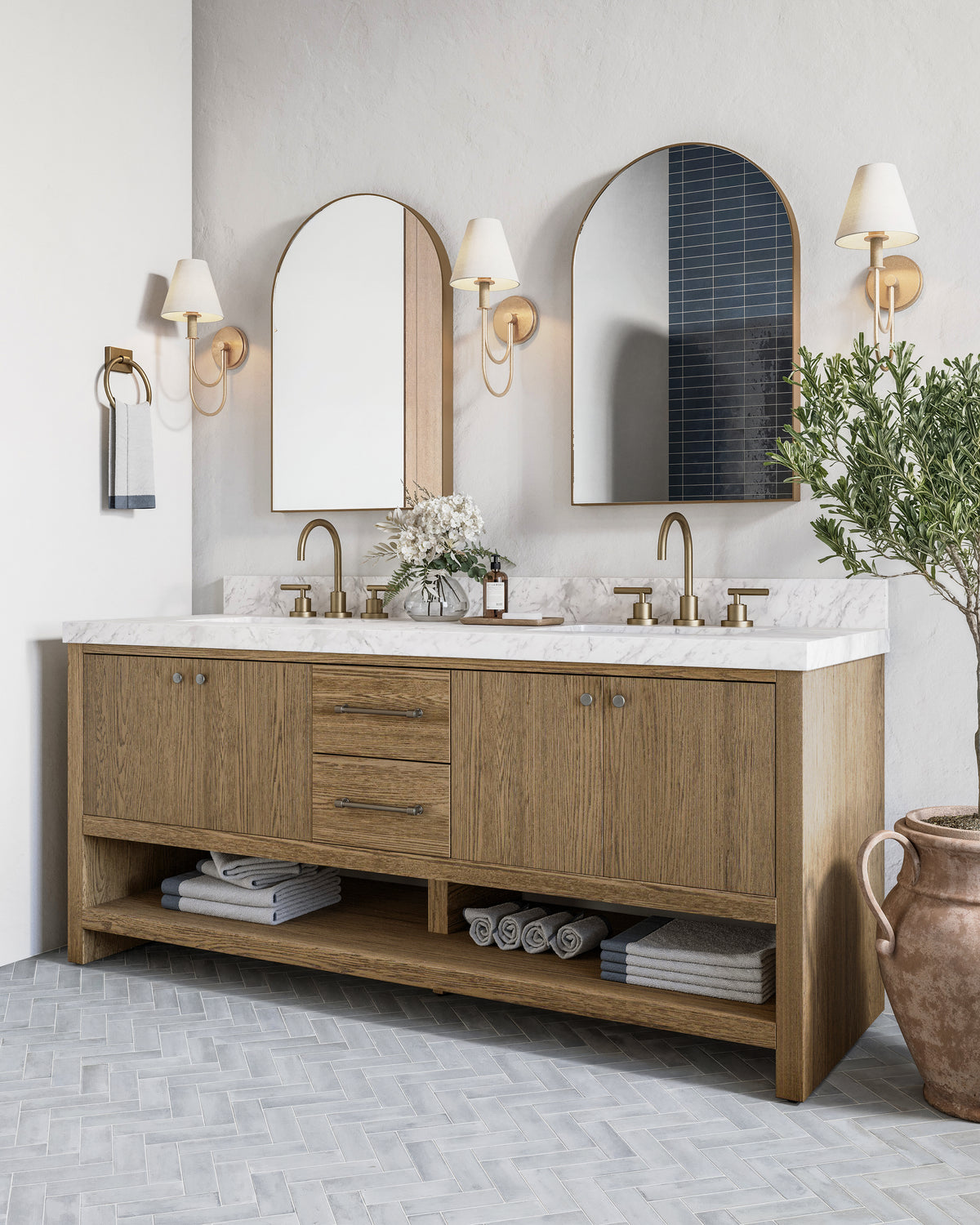Elegant bathroom with wood double vanity, marble countertop, arched mirrors, brass sconces, and gray herringbone tile floor.