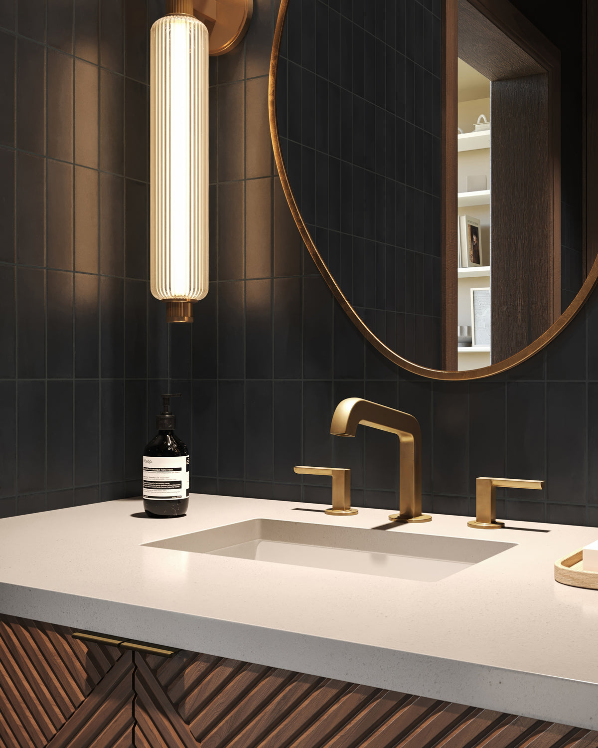 Antique brass wall sconce with a vertically ribbed glass cylinder casting warm ambient light beside a round brass mirror in a modern bathroom with dark tile walls.
