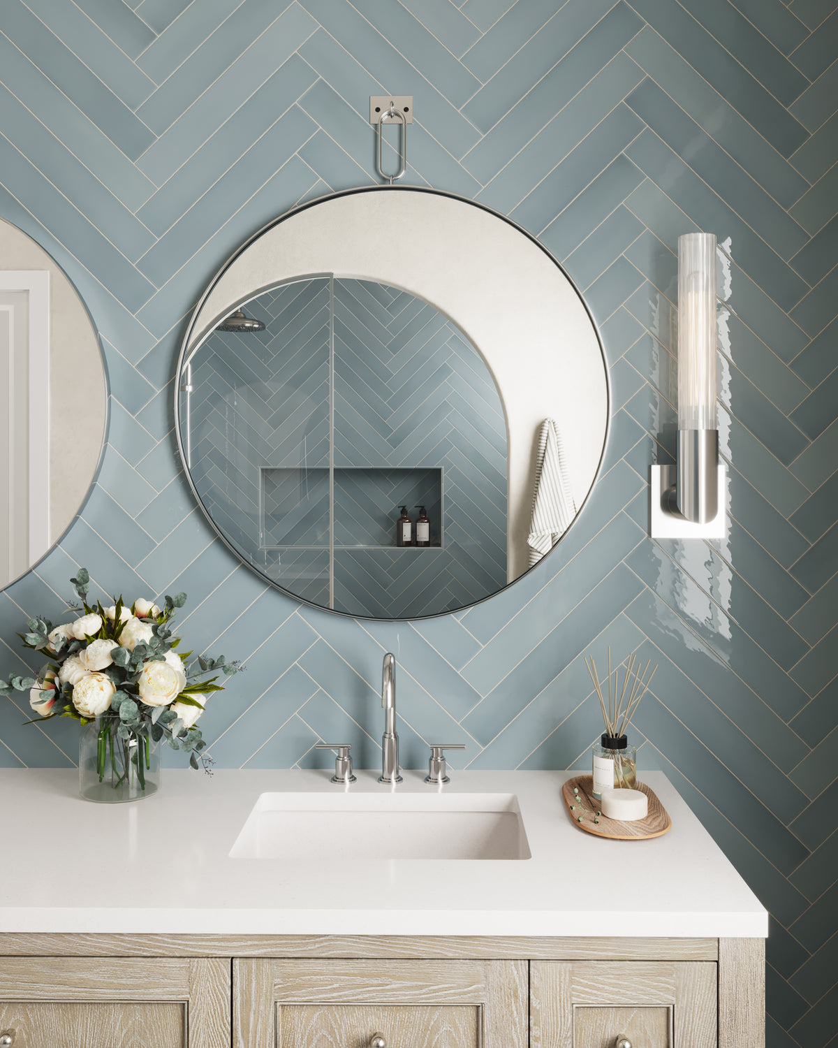 Whitewashed oak bathroom vanity with paneled drawers and round knobs, topped with a white quartz countertop and integrated sink. A large round mirror and vertical glass wall sconce are mounted on a blue herringbone tile wall, with a vase of flowers and small tray arranged on the counter.