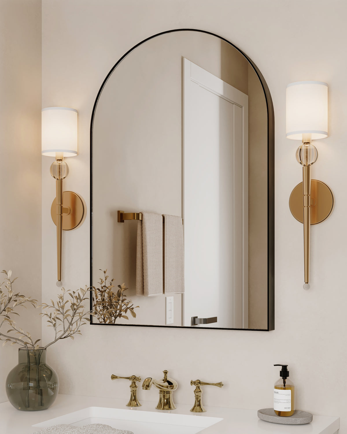 Brass and crystal wall sconces with white fabric shades frame an arched mirror above a vanity with brass fixtures in a serene, neutral-toned bathroom