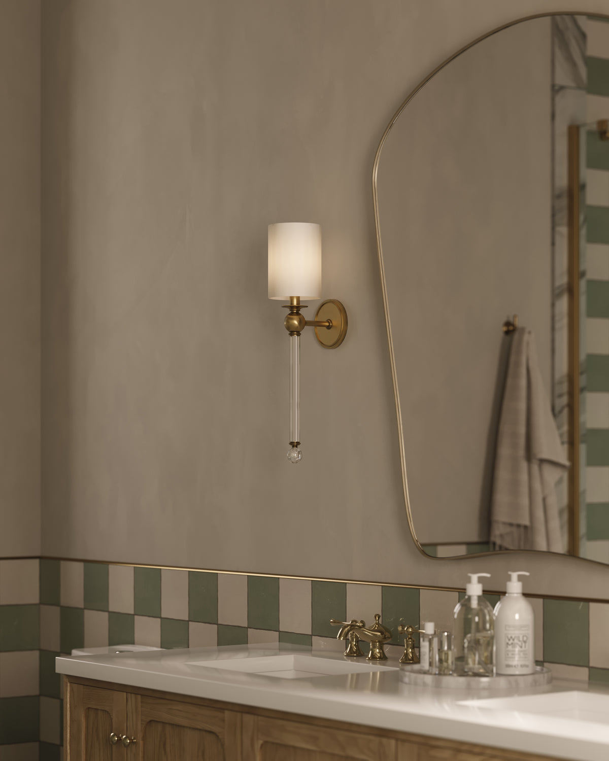 Elegant aged brass wall sconce with white shade and crystal rod detail illuminating a vanity with wood cabinet and checker tile backsplash.