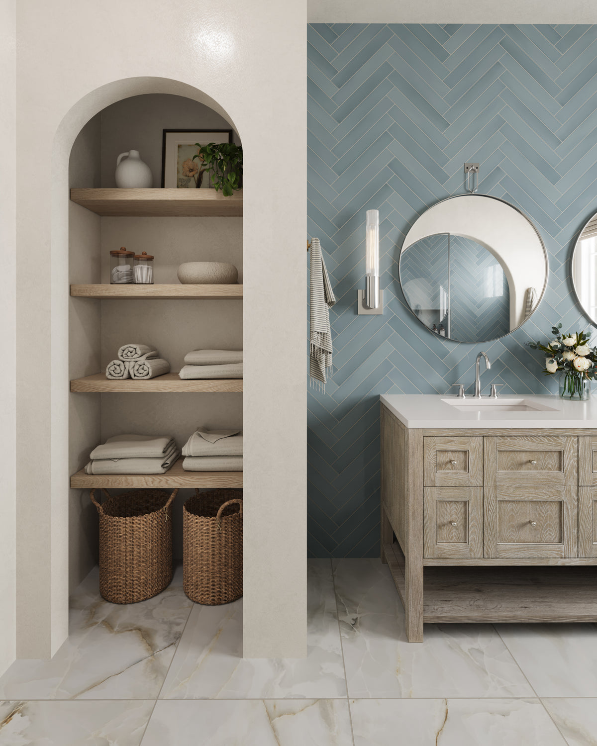 Bathroom with a whitewashed oak vanity and white quartz countertop set against a blue herringbone tile wall with round mirror and glass sconce. Beside it, an arched built-in shelving niche with wooden shelves neatly holds folded towels, jars, decor, and woven baskets on a marble floor.