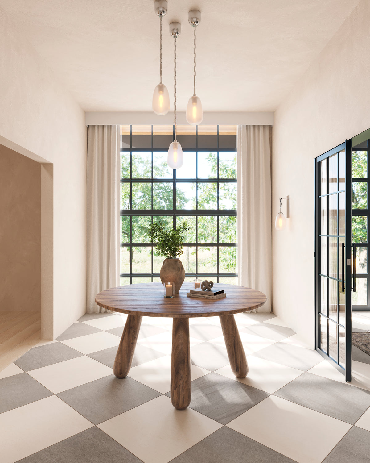 Round wood table with chunky sculptural legs sits atop a checkerboard tile floor, framed by arched windows and soft pendant lighting.
