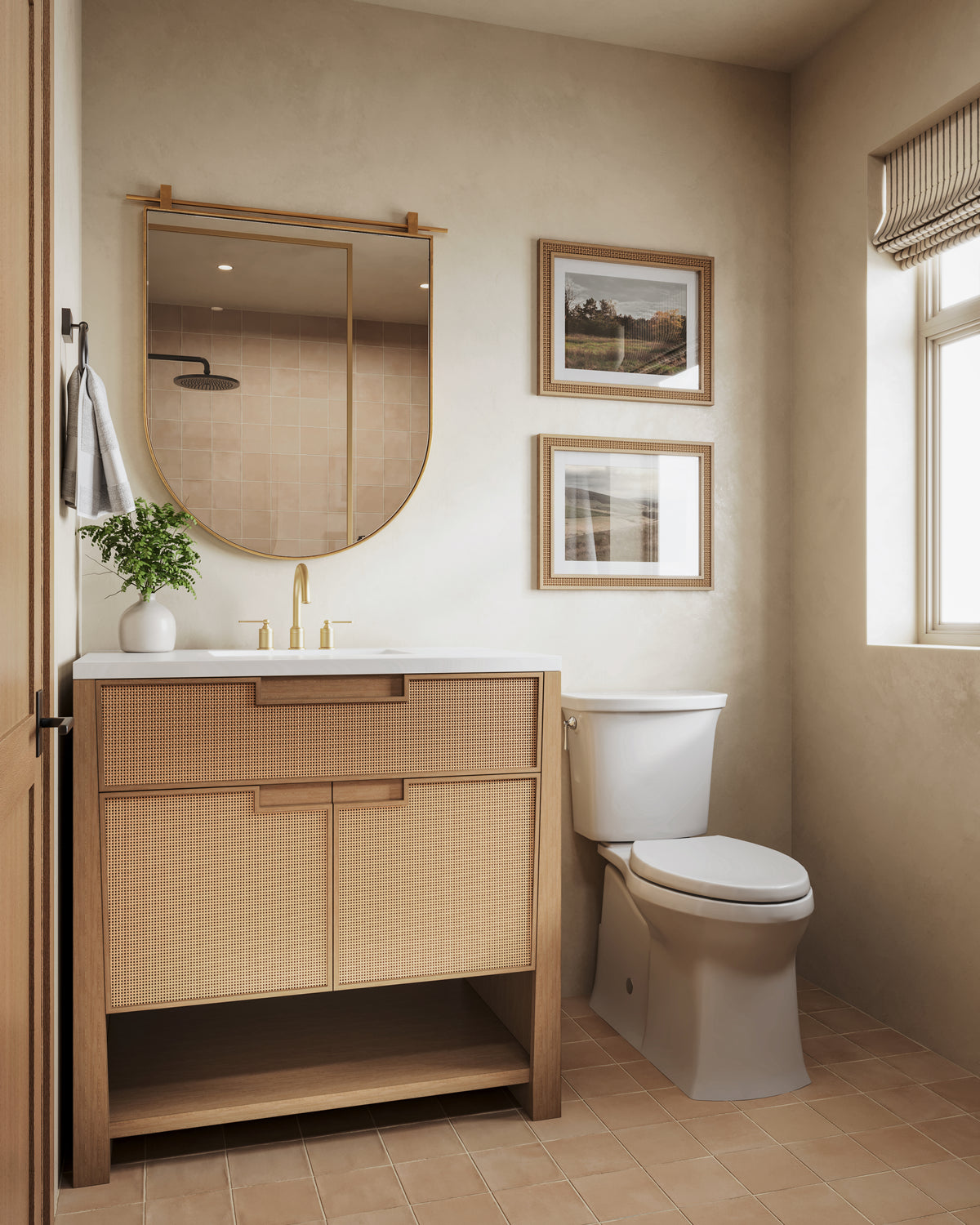 Compact bathroom with oak cane vanity, brass faucet, arched mirror, framed art, toilet, and warm beige wall tiles with natural light.
