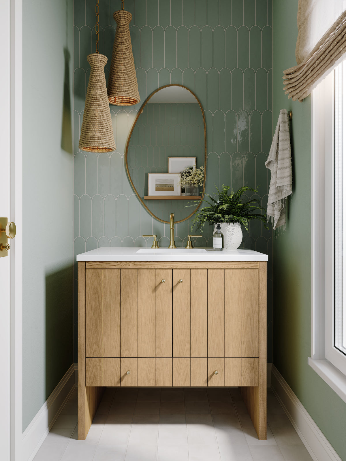 Light natural oak bathroom vanity with vertical wood grain doors and brass knobs, topped with a white quartz countertop and brass faucet. An oval brass-framed mirror hangs above sage green scalloped tiles, with woven pendant lights, a small plant, and a window bringing soft natural light into the space.