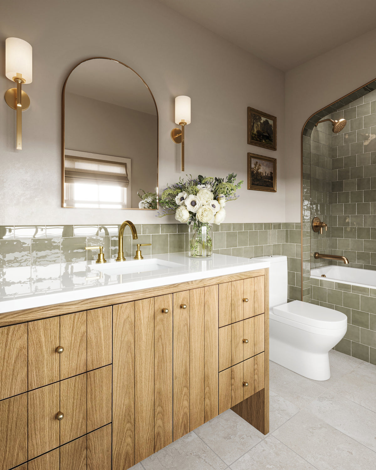Light natural oak bathroom vanity with vertical wood grain doors and side drawers with brass knobs, topped with a white quartz countertop and brass faucet. An arched mirror and brass wall sconces hang above soft green tile wainscoting, while a glass vase of white flowers sits beside the sink and an arched glass shower with matching green tiles appears in the background.