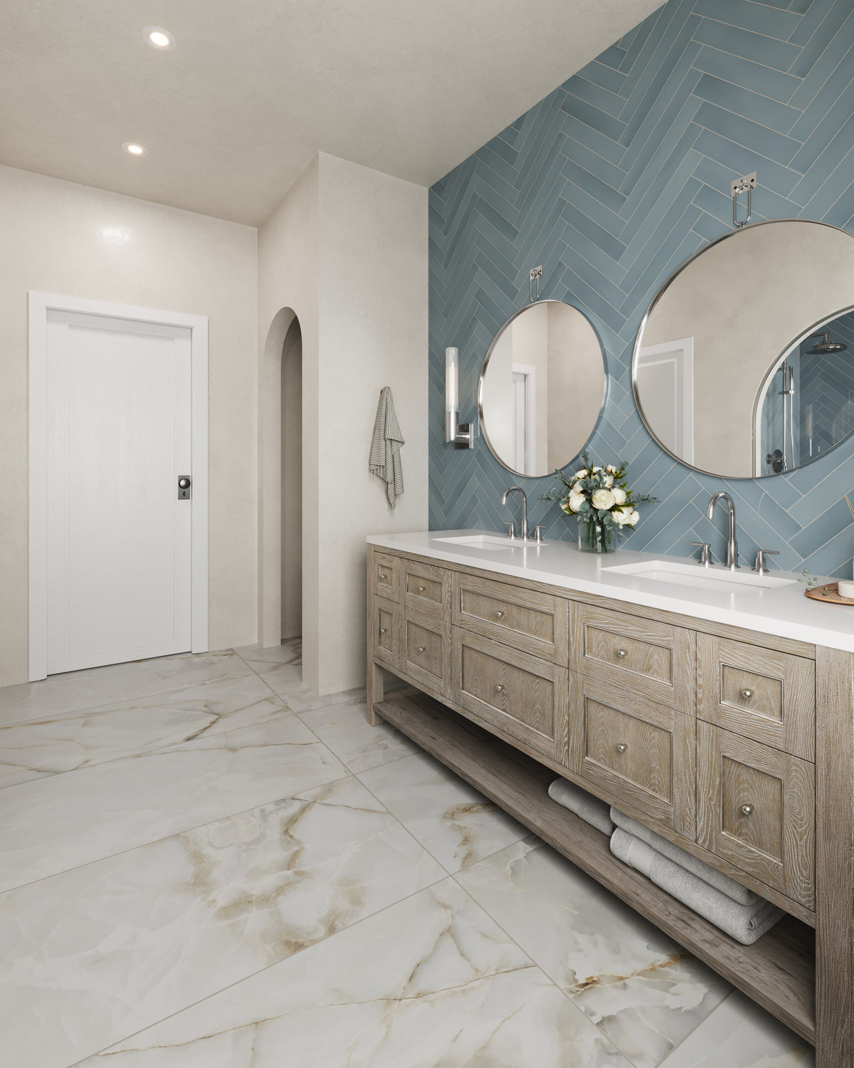 Spacious bathroom with a whitewashed oak double vanity featuring paneled drawers and an open lower shelf, topped with a white quartz countertop and dual sinks. Two round mirrors and glass wall sconces hang against a blue herringbone tile accent wall, while large marble-look floor tiles and an arched wall niche complete the bright, modern space.