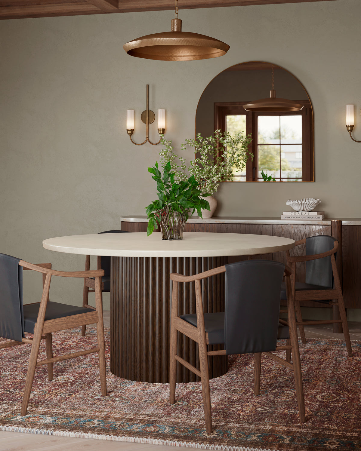 Round dining table with sculptural wood base and leather chairs, styled with greenery, brass lighting, and layered textures for a warm dining space.