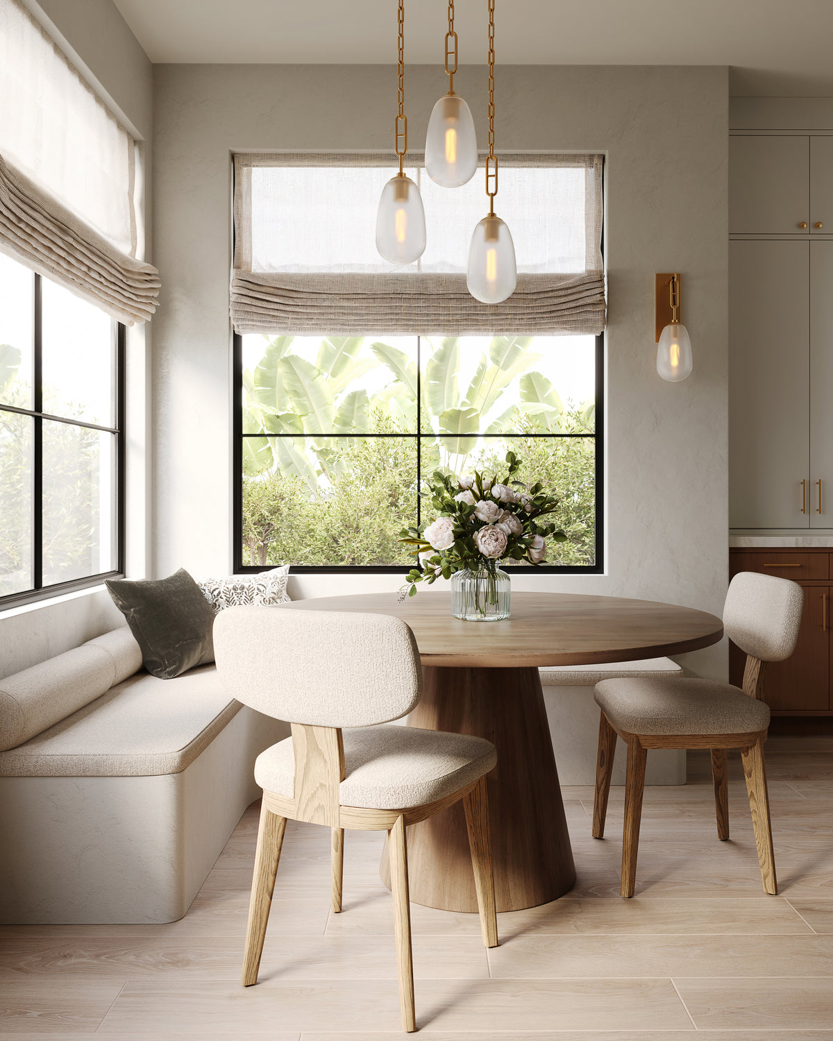 Cozy breakfast nook with round wooden table, upholstered seating, layered pendant lights, and greenery framed by large windows.