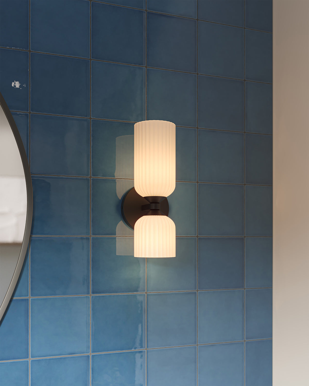 Wall vanity light with a dark bronze mount and ribbed glass shades installed on glossy blue wall tiles beside a mirror, casting soft warm light in a modern bathroom.