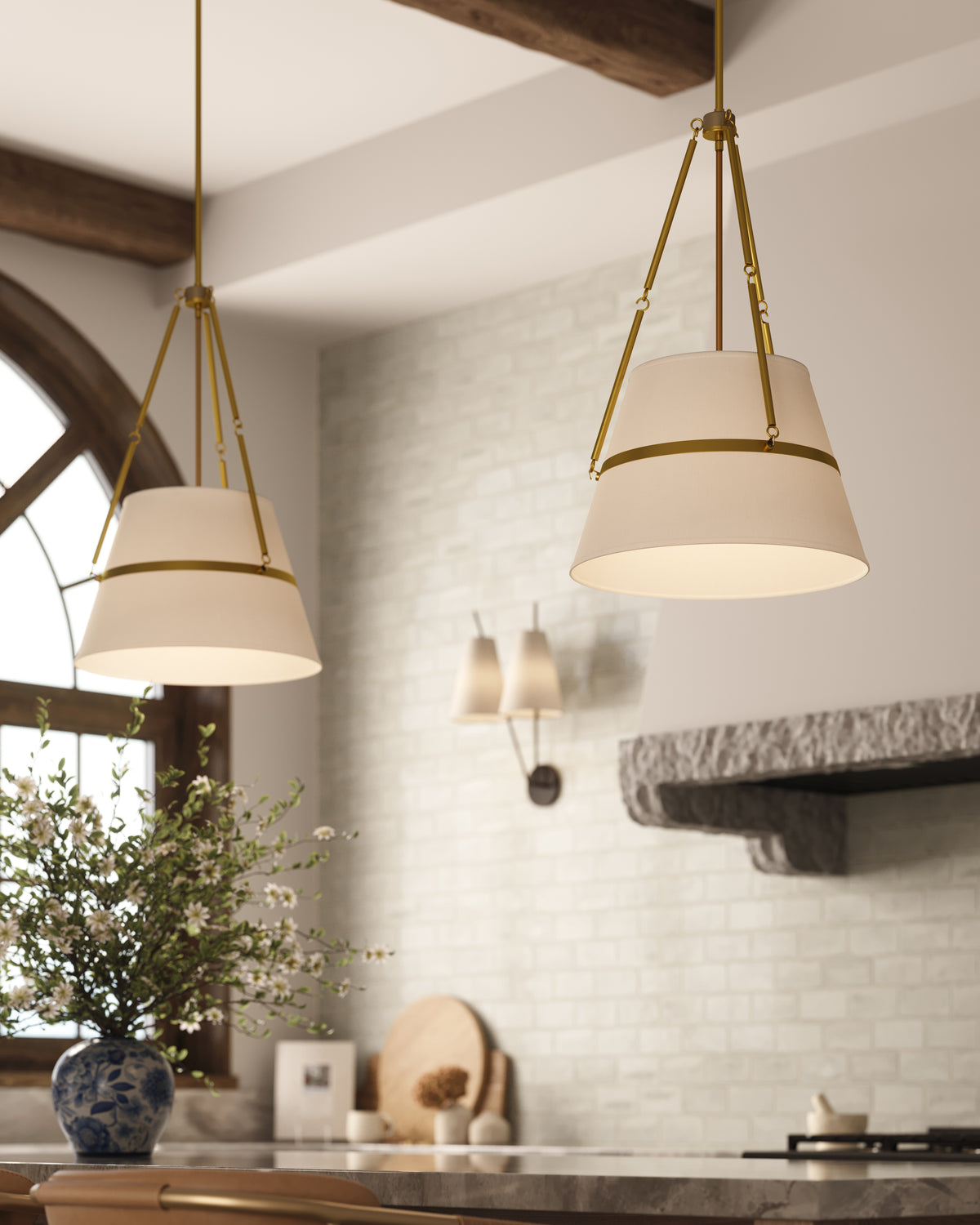 Pair of brushed gold pendant lights with tapered white fabric shades suspended above a kitchen counter with white brick walls and rustic wood beams.