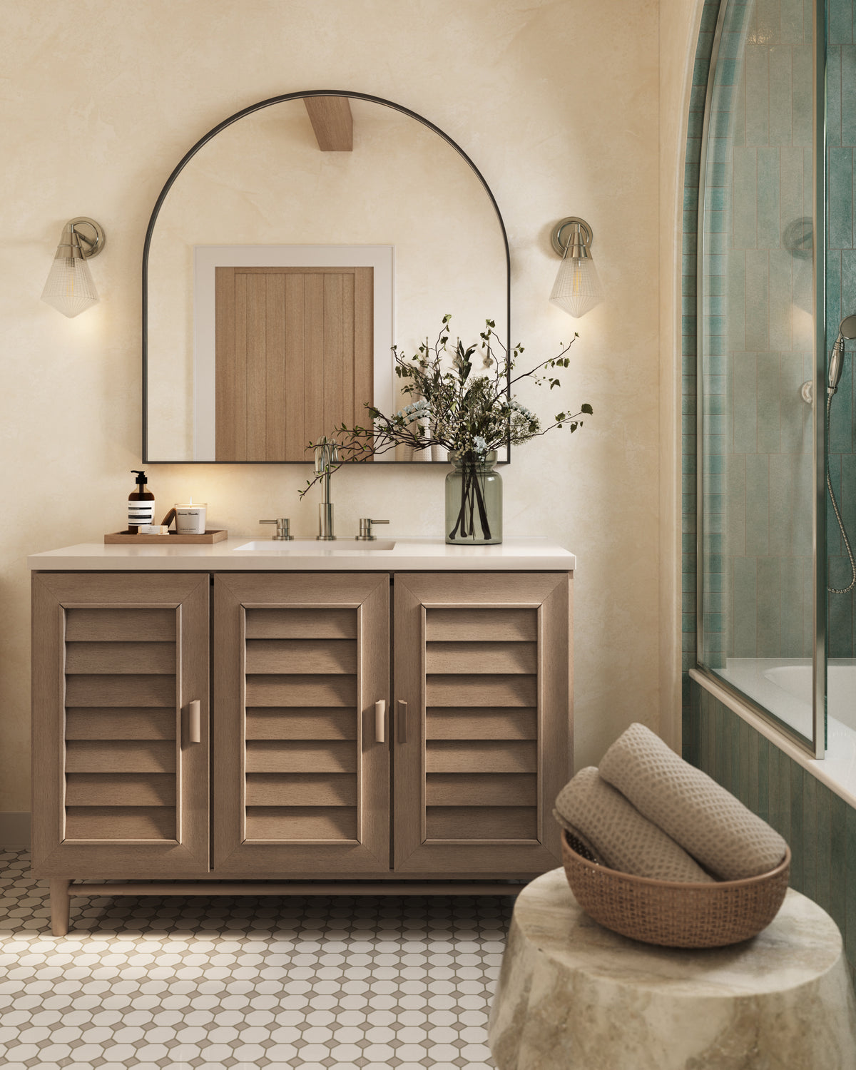 Whitewashed walnut bathroom vanity with louvered cabinet doors, white quartz countertop, archedmirror, wall sconces, and floral arrangement beside a glass shower in a warm neutral bathroom.