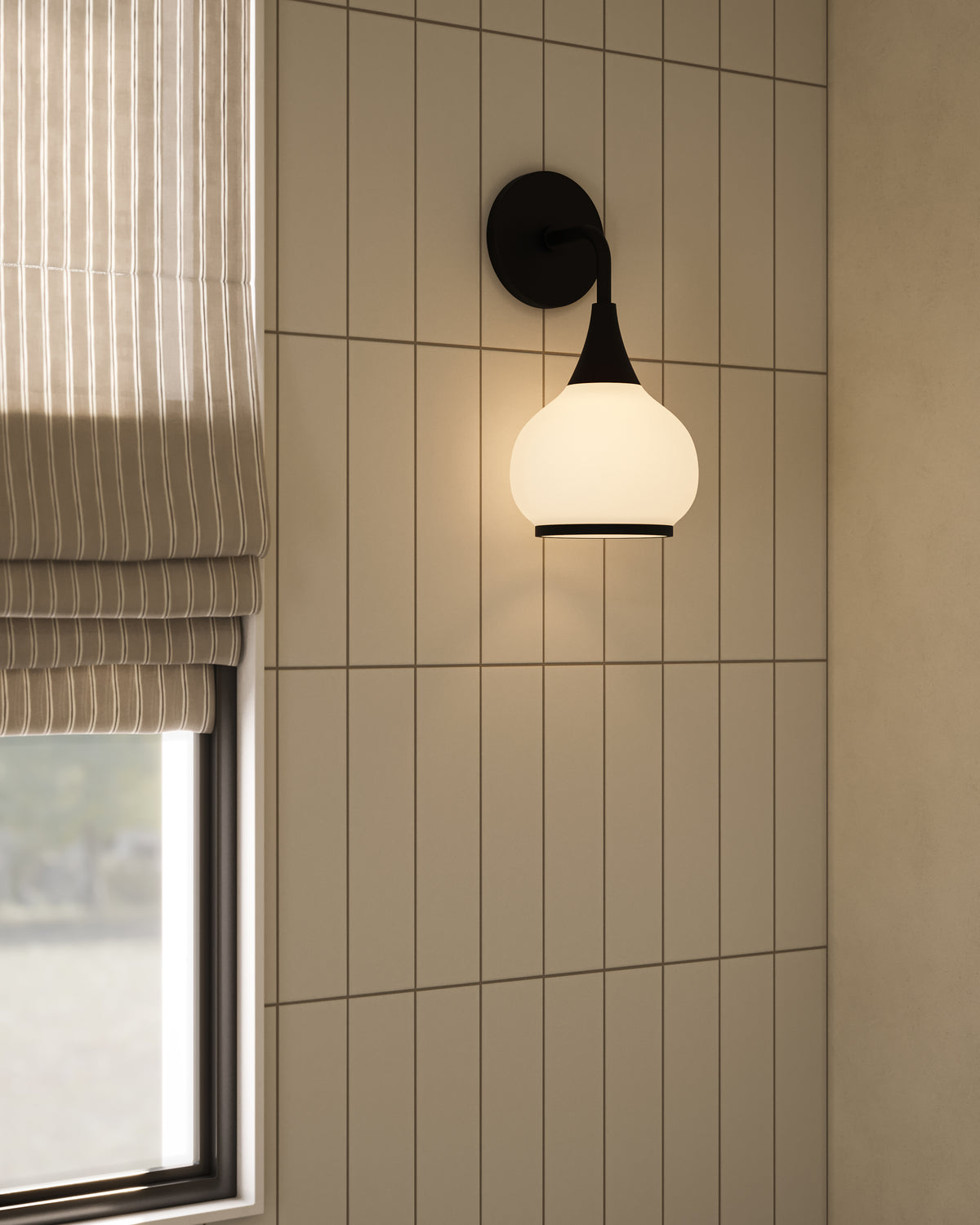 Matte black wall sconce with opal glass globe shade mounted on a vertically tiled wall beside a window with a striped Roman shade.