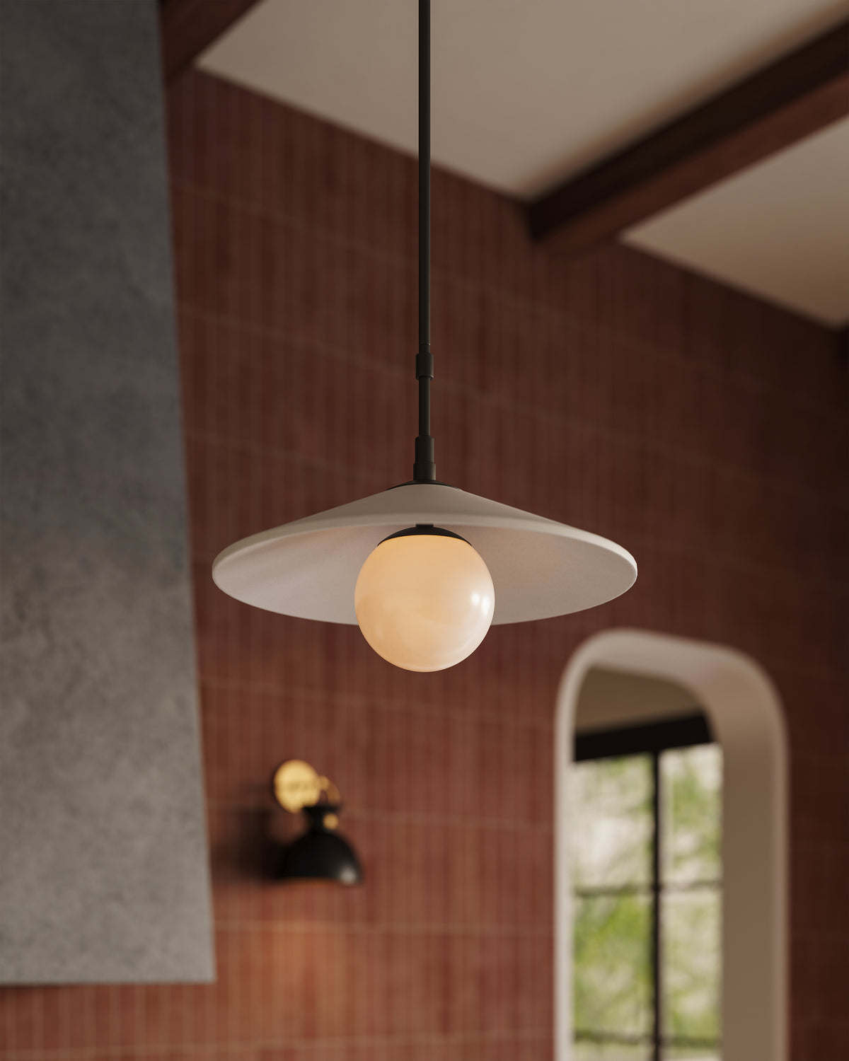 Modern pendant light with black extendable stem, beige shade, and glowing globe bulb over terracotta subway tile wall.