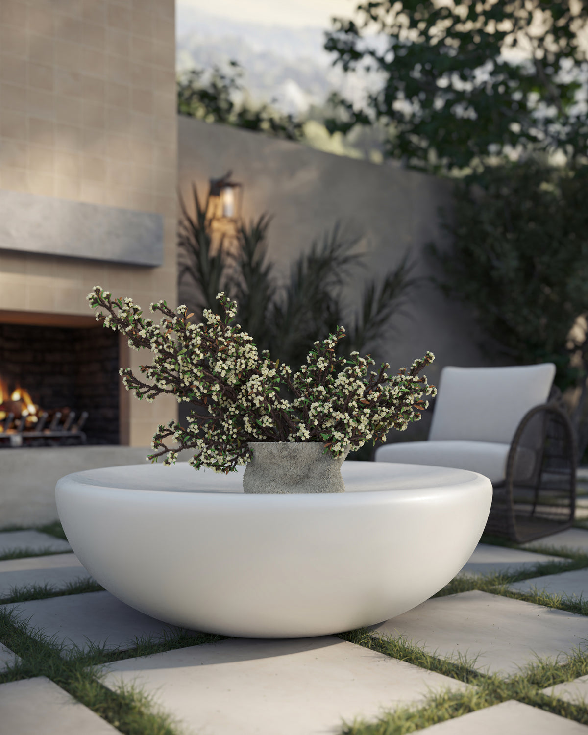 Outdoor patio with a white round concrete coffee table holding a stone planter of greenery, set on large square pavers with grass joints near a lit fireplace, lounge seating, garden plants, and mountain views in the background.