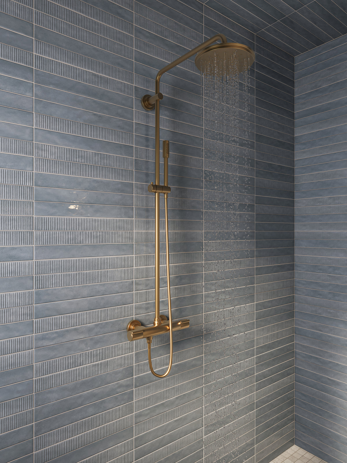 Walk-in shower with blue-gray ribbed wall tiles, brass rainfall showerhead, handheld shower fixture, and water flowing against the tiled wall.
