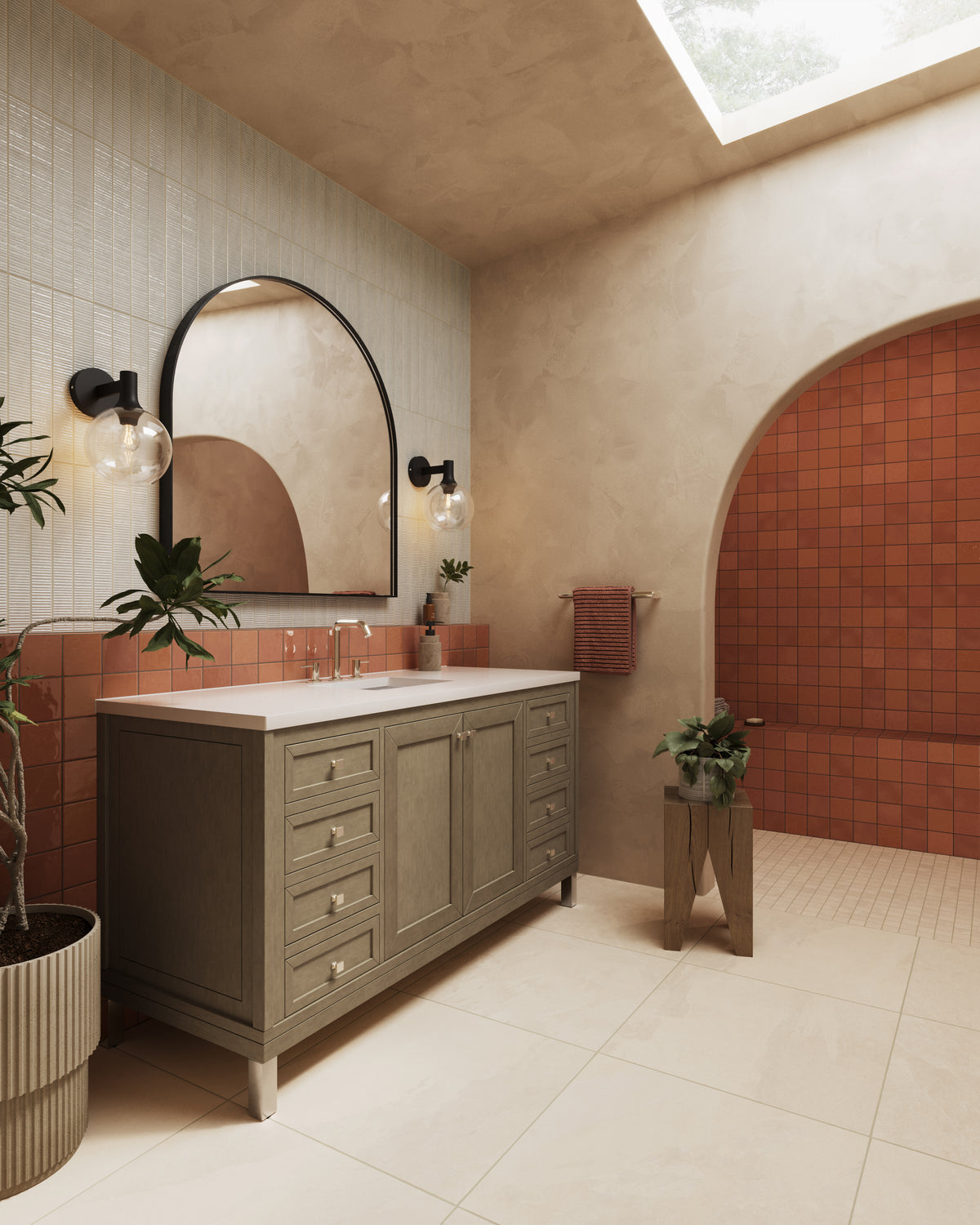Warm bathroom vanity scene with glossy rust square tile used as a backsplash and shower accent, paired with cream reeded wall tile, beige plaster walls, an arched mirror, black sconces, and a skylight-lit neutral floor.