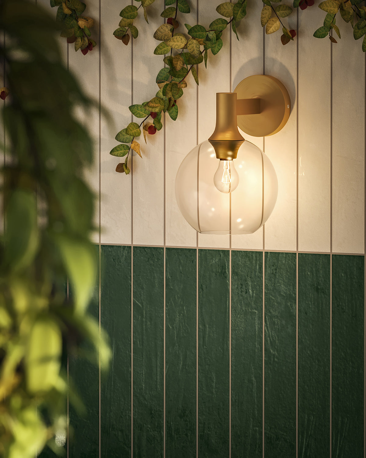 Modern wall sconce with clear glass globe shade and brushed brass mount on green and white subway tile wall.