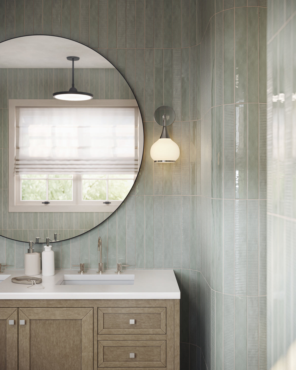 Bathroom vanity with pale green vertical wall tiles, large round black-framed mirror, globe wall sconce, and light wood cabinetry.