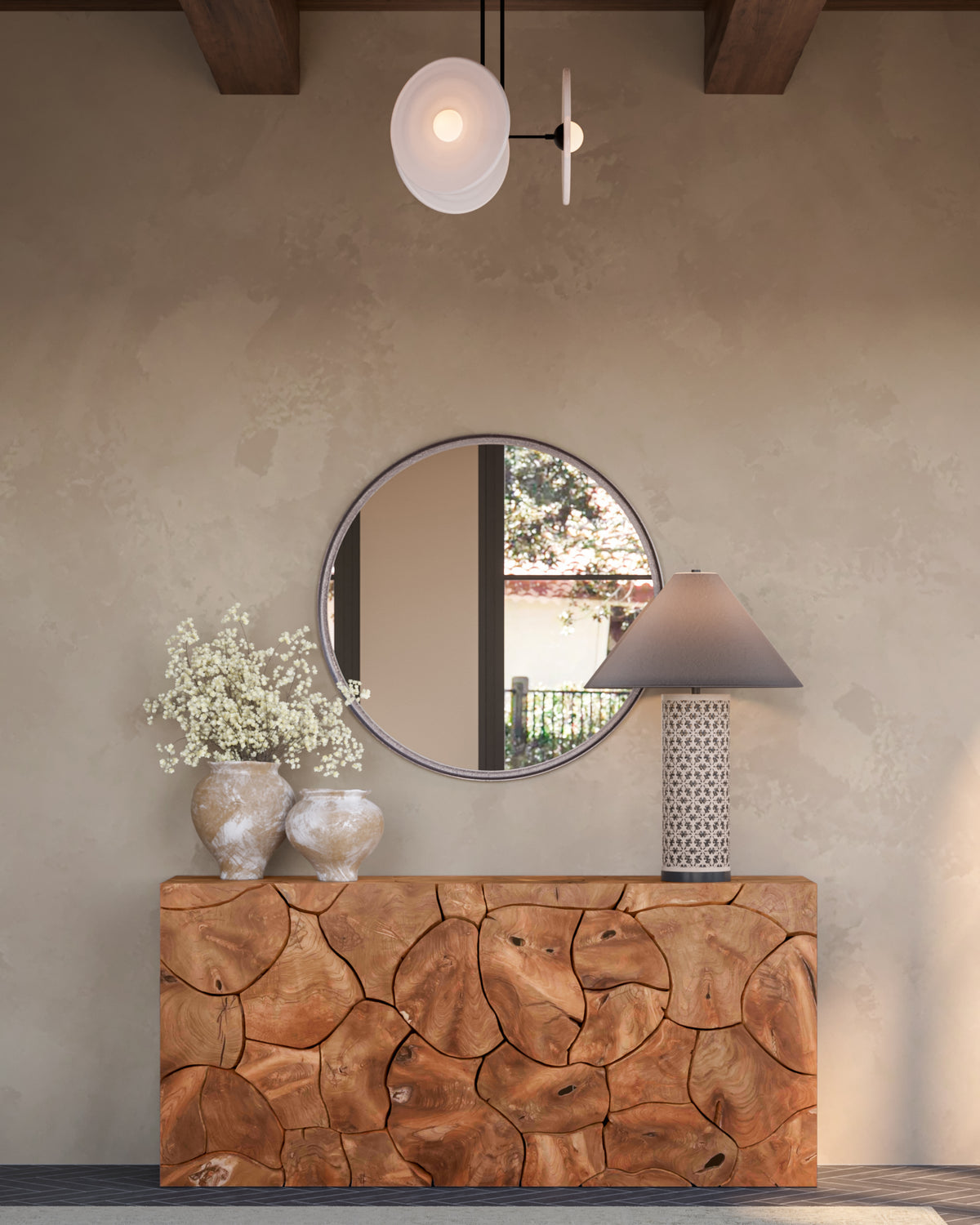 Warm entryway with a cream and black patterned table lamp, dark tapered shade, carved wood console, round mirror, white ceramic vases, dark herringbone floor tile, and textured plaster wall.