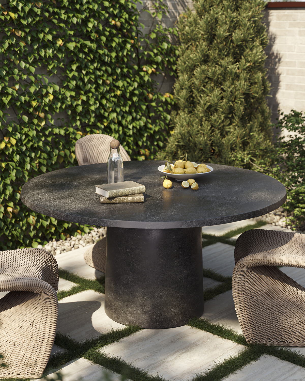 Outdoor garden patio with a round aged dark gray pedestal dining table set on square pavers with grass joints, surrounded by woven lounge chairs, ivy-covered walls, greenery, and a tabletop arrangement of lemons, books, and a glass bottle.