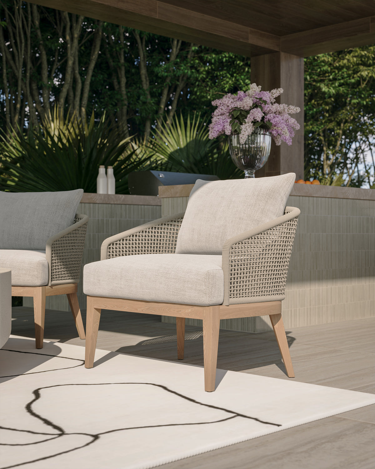 Cream cushioned outdoor lounge chair with beige woven rope arms and a light wood frame styled on a covered patio with pale floor tile, a white rug, tropical greenery, and a tiled outdoor bar.