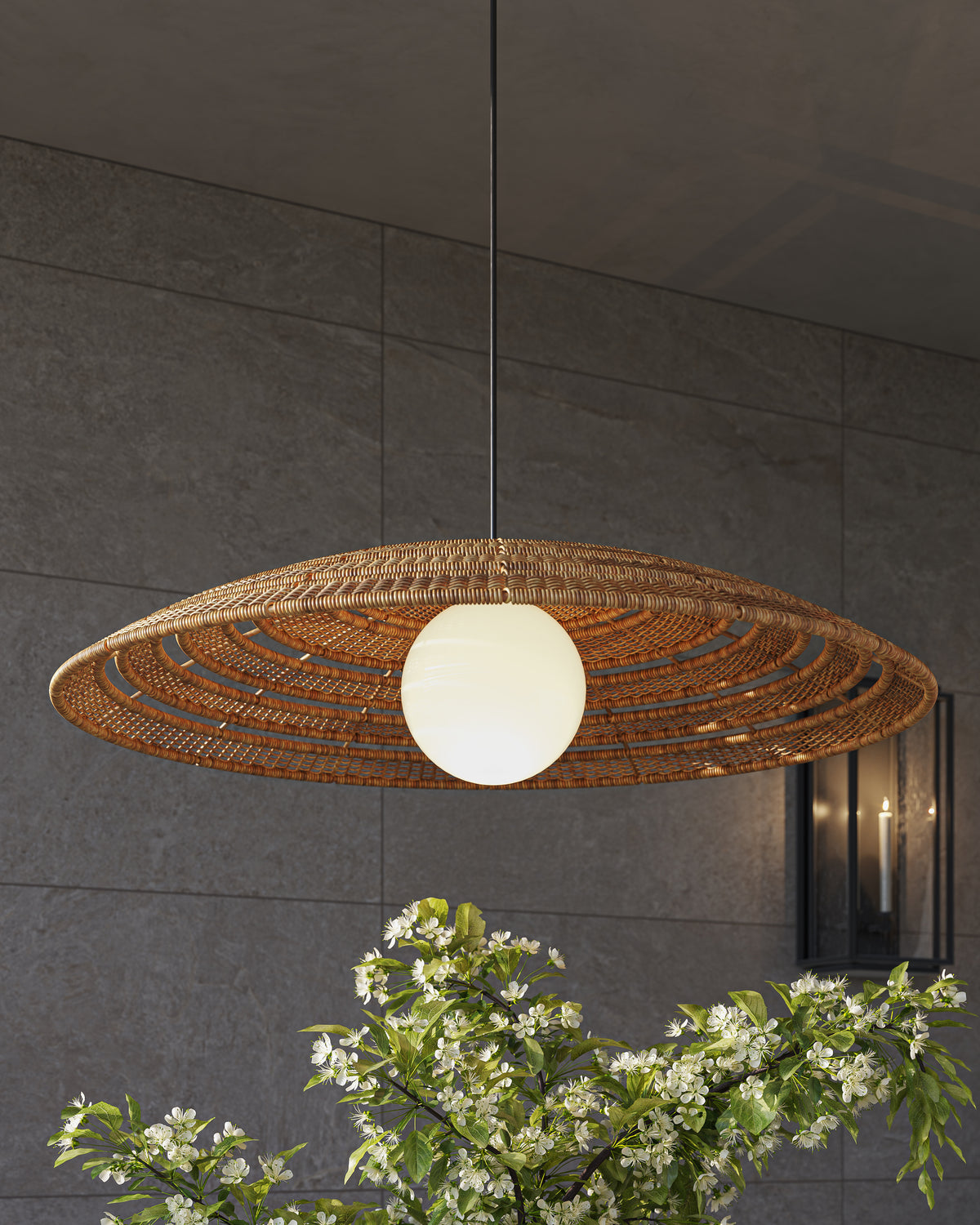 Our Rivington Outdoor Pendant Large with a wide flat honey brown wicker dish shade and white milk glass center, hanging over green plants against a dark gray stone wall tiles.