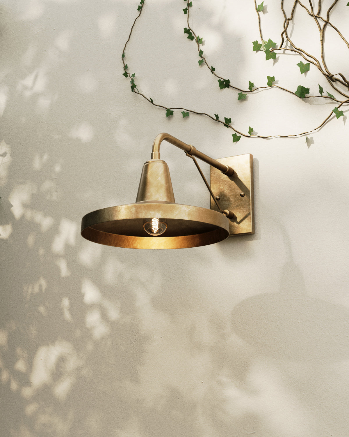 Antique brass outdoor wall sconce mounted on a stucco wall, featuring an angled arm, rectangular backplate, umbrella-style shade, and exposed bulb beneath trailing ivy and soft leaf shadows.