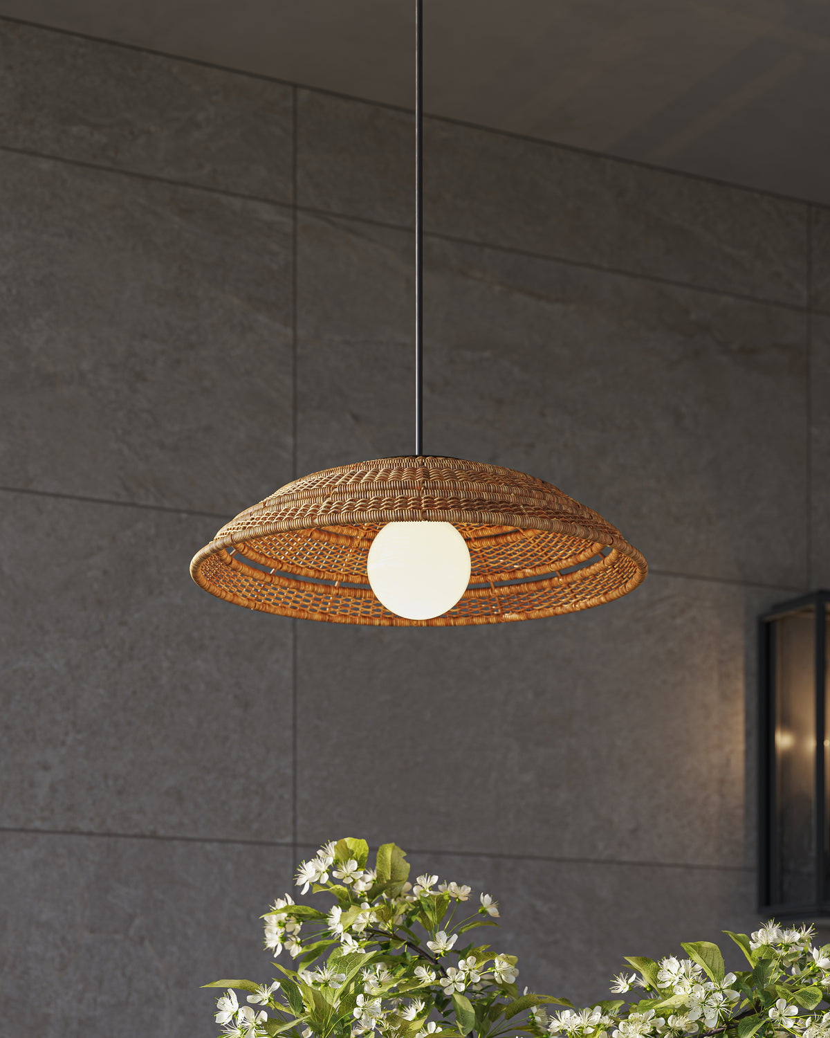 A compact shallow honey brown wicker dish pendant with a glowing white round center light, suspended outdoors against a dark gray textured stone wall tile with white blooms below.

