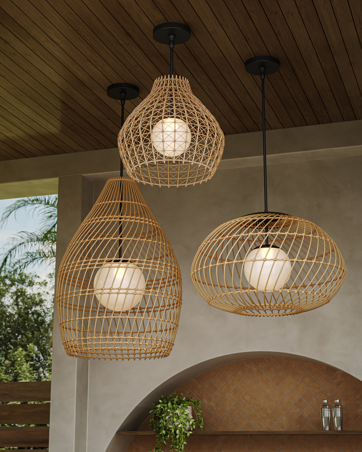 Three honey brown wicker open-weave pendants with glowing white centers in teardrop and dome silhouettes, suspended from a dark wood ceiling on a warm outdoor covered patio.