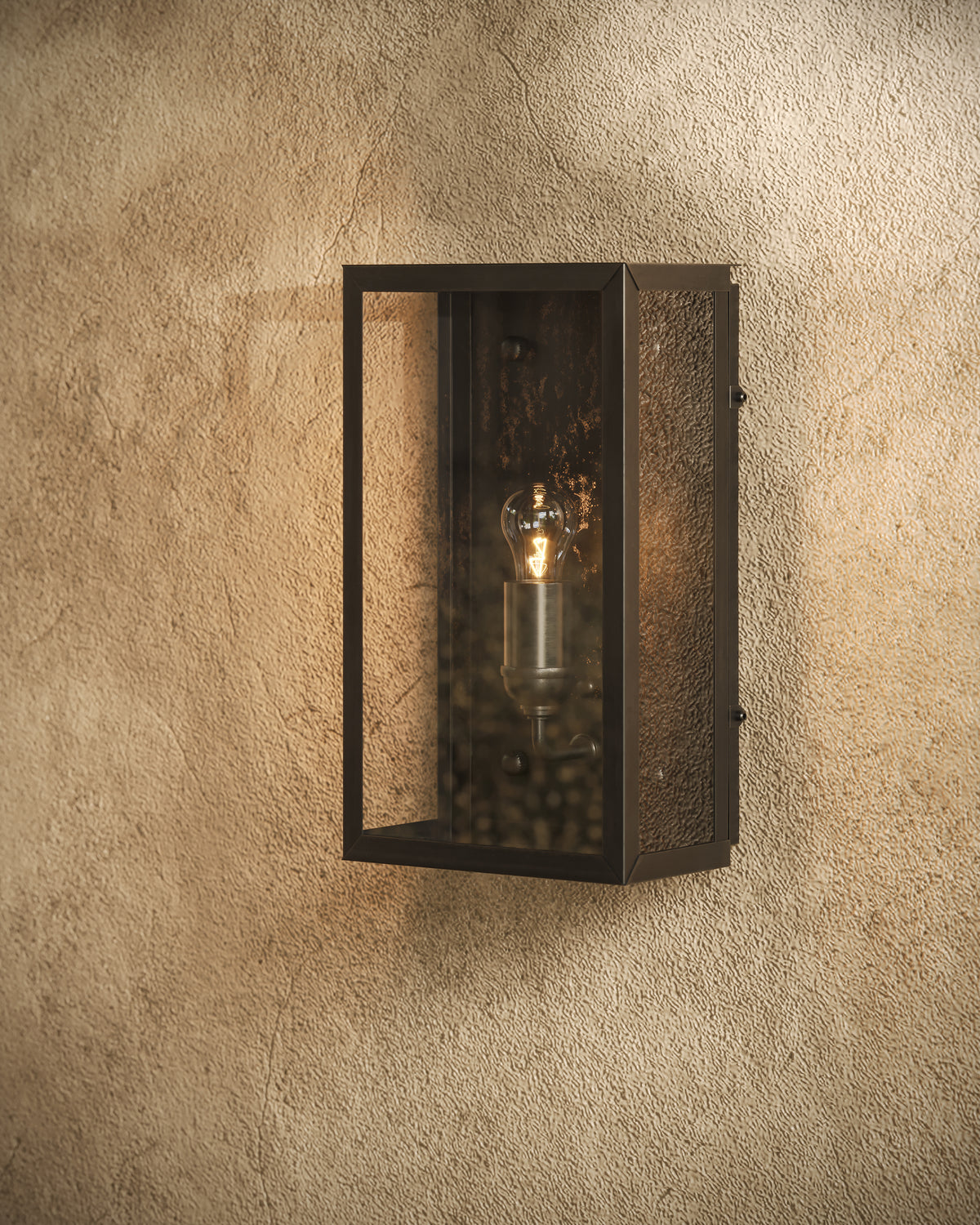 Matte black wall lantern with clear glass panels and a glowing exposed bulb mounted on a textured beige stucco wall, casting warm light across the surface.