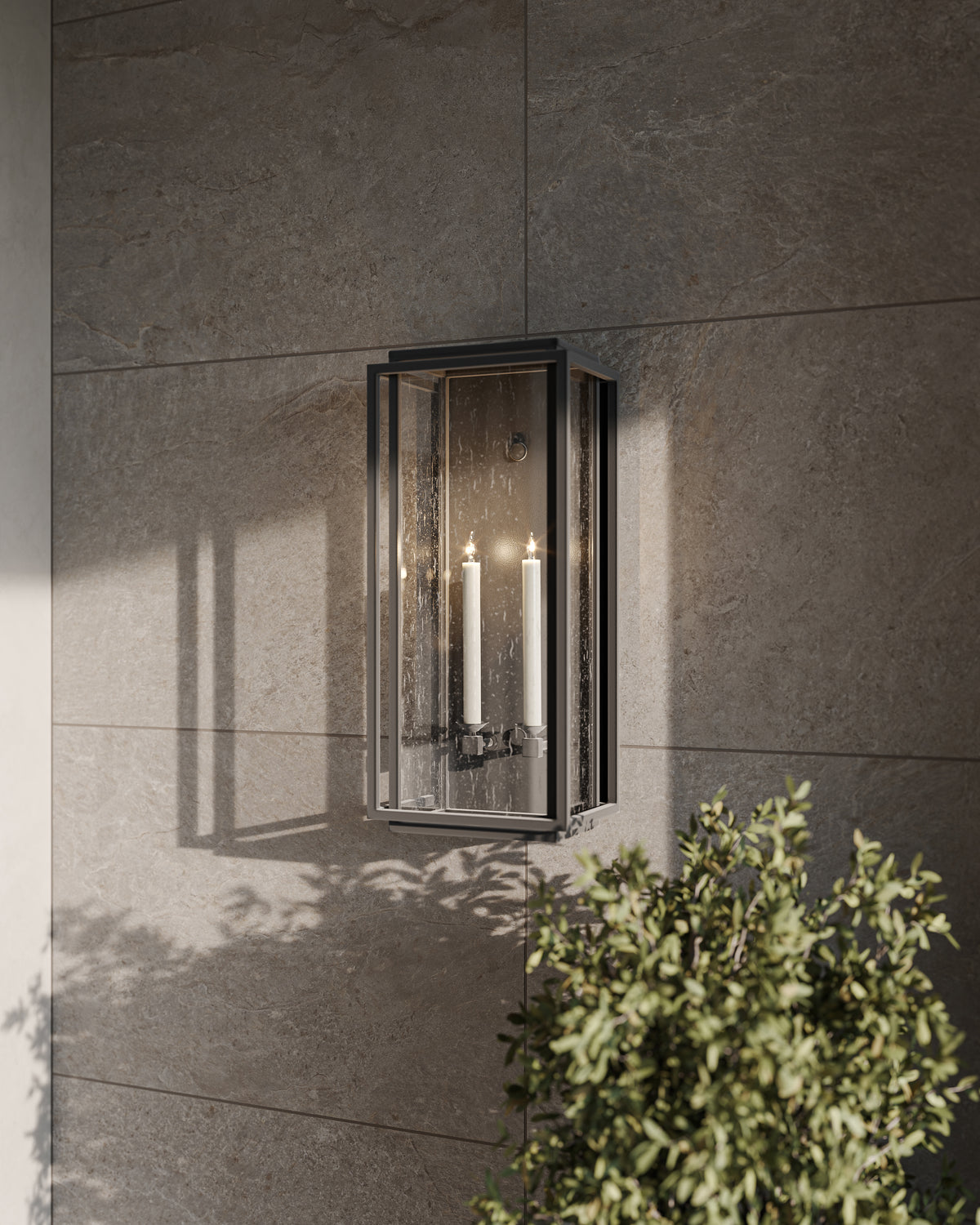 Black glass wall sconce with two candle-style lights mounted on a gray stone tile wall beside green outdoor foliage.