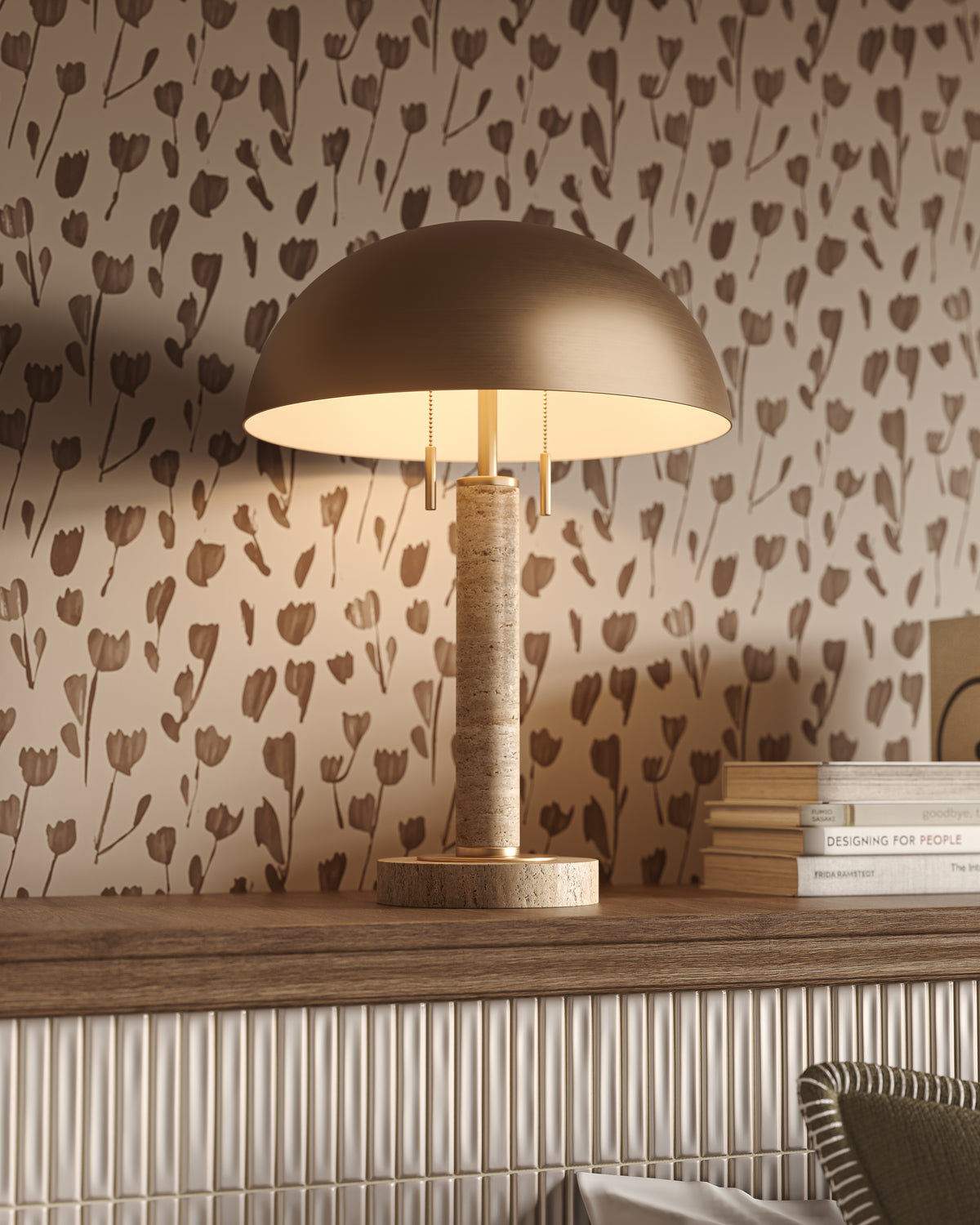 Travertine table lamp with warm brass semi-spherical metal shade, twin pull chains, and soft floral wallpaper behind.