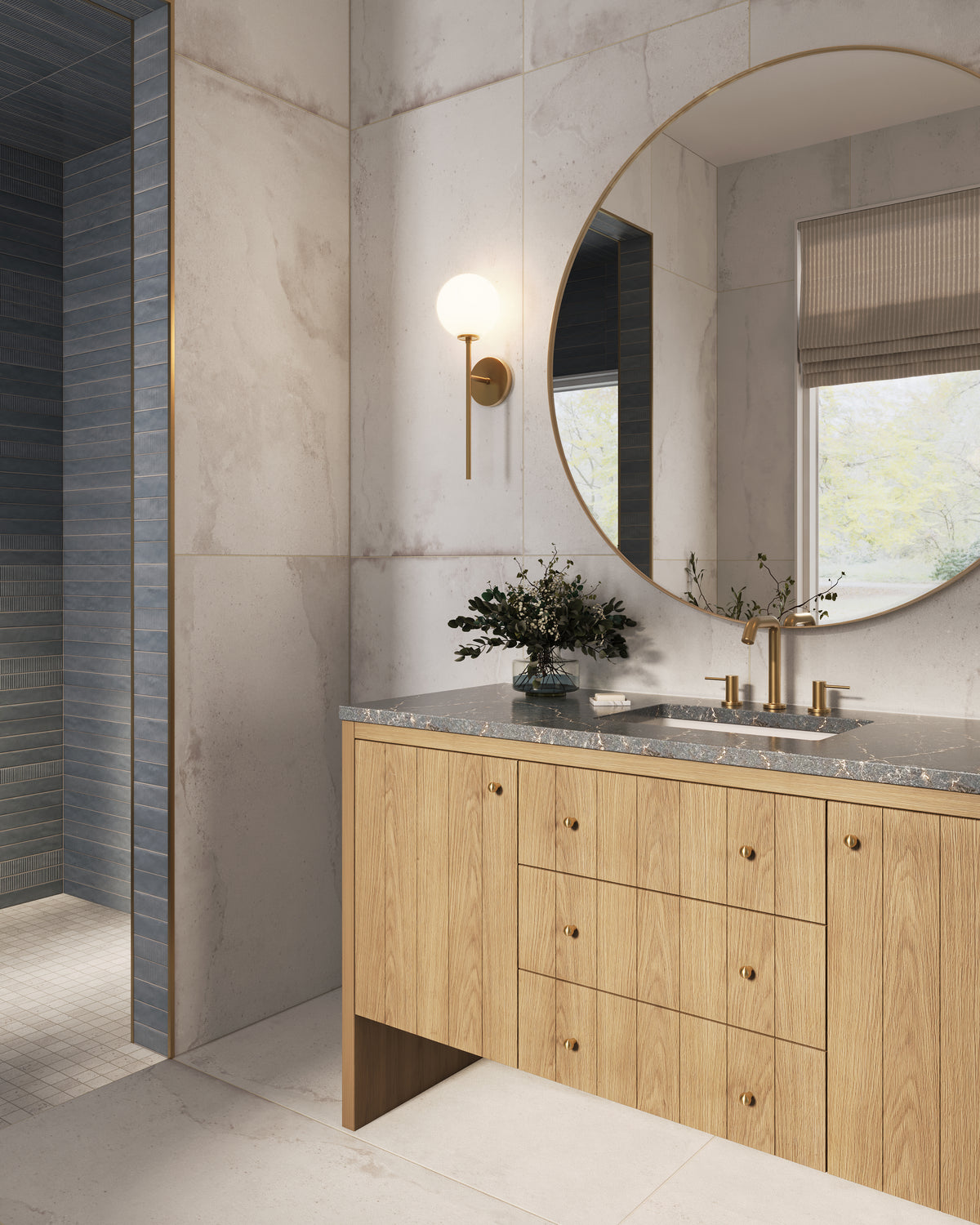 Bathroom vanity area with large marble-look wall tile, a light wood vanity topped with gray stone, brass fixtures, a round mirror, and a nearby walk-in shower with blue stacked subway wall tile and off-white matte mosaic floor tile.